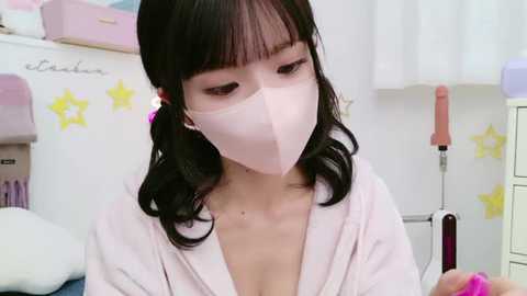 Media: Video of an East Asian woman with pale skin, long black hair, and bangs, wearing a pink mask, light robe, and pink earrings. She is in a pastel-colored, star-decorated room with a white shelf and a pink stool.