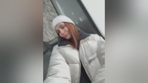 Media: A video of a Caucasian woman with long red hair, wearing a white beanie and a puffy white jacket, standing in a dimly lit, gray hallway with textured walls.