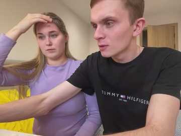 Media: Video of a blonde woman in a purple shirt and a young man in a black Tommy Hilfiger shirt, both looking serious in a beige room with a yellow blanket.