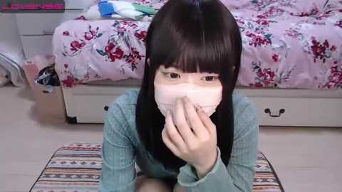 Media: Video of an Asian woman with straight black hair, wearing a face mask, kneeling on a patterned rug in a bedroom with a floral bedspread.