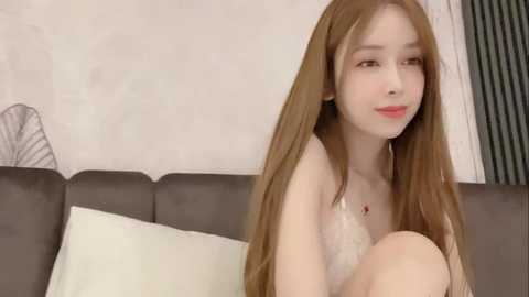 Media: Video of a fair-skinned, young Asian woman with long, straight brown hair, sitting on a gray couch. She wears a white bralette, and the background features a white wall and a striped window.