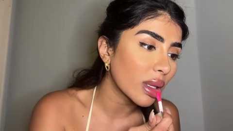 Media: Video of a young, light-skinned woman with long, dark hair, wearing a gold necklace, applying pink lipstick with a brush, against a neutral background.