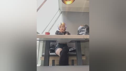 Media: Video of a young woman with short blonde hair, wearing a black outfit and headphones, sitting at a modern, glass-top table in a minimalist, well-lit room.