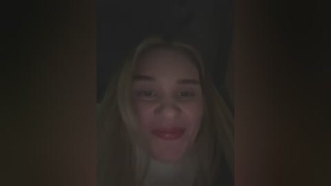 Media: A dimly lit video of a woman with long, wavy blonde hair, fair skin, and red lipstick, smiling directly at the camera, with a dark background.