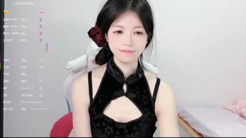 Media: Video of a young Asian woman with fair skin, black hair, and a red flower accessory, wearing a black velvet dress with a high collar, seated indoors with a digital interface in the background.