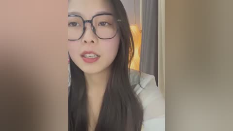 Media: Video of an Asian woman with long black hair, wearing large black-framed glasses and a white shirt, captured indoors with soft lighting.