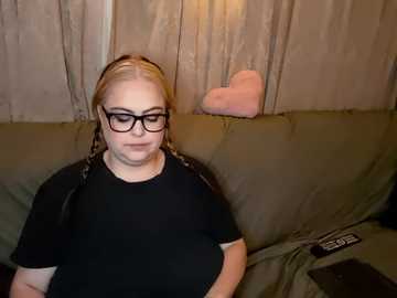 Media: Video of a fair-skinned, blonde woman with glasses, wearing a black top, sitting on a green couch with beige curtains in the background.