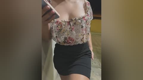 Media: Video of a woman in a floral lace top and black mini skirt, taken indoors with a yellow wall and white sheet in the background.