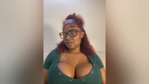 Media: Video of a dark-skinned Black woman with curly, maroon hair, wearing black-framed glasses, a low-cut green top revealing ample cleavage, and a gold necklace. She stands against a plain beige wall.