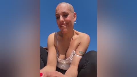 Media: A video of a smiling, bald woman with light skin, wearing a white lace bra and black pants, sitting on a motorcycle against a clear blue sky background.