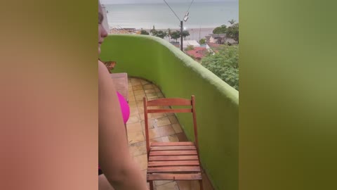 Media: A video of a woman in a pink bikini standing on a green-painted balcony, with a wooden chair in the foreground and a scenic view of a coastal town and ocean in the background.