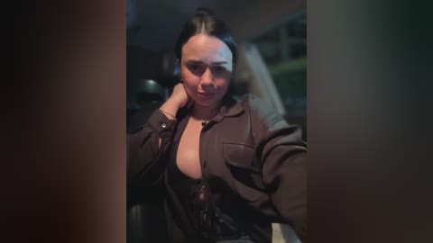 Media: A video of a young woman with fair skin, dark hair, and a medium build, wearing a low-cut brown jacket, leaning against a dark car interior.