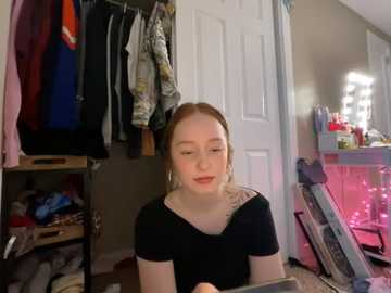 Media: Video of a young Caucasian woman with light skin and straight, blonde hair, wearing a black top, sitting in a cluttered bedroom with a closet full of clothes, makeup table, and pink fairy lights.