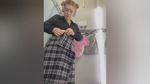 Media: Video of a young, fair-skinned woman with red hair styled in buns, wearing a black blouse and a black-and-white plaid skirt, adjusting her outfit in a modern, minimalist room with white walls and a pink garment hanging on a hook.