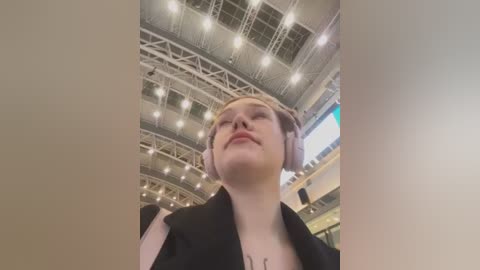 Media: Video of a young woman with short blonde hair, wearing a black jacket, standing inside a large, modern building with a high ceiling, visible structural beams, and bright lighting.