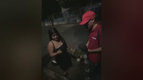 Media: Video: A young Latina woman with long dark hair and a black tank top, holding a green object, talks to a red-shirted man with a cap in a dimly lit, urban setting.