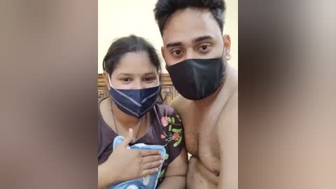 Media: A video captures a young South Asian couple, both wearing masks, indoors. The woman, with medium skin tone and dark hair, smiles. The man, with a beard and short black hair, is shirtless, showing his muscular physique.