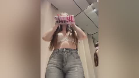 Media: Video of a young woman with long, light brown hair, wearing a white lace bra and high-waisted, light-wash jeans, taking a selfie in a fitting room, reflected in a full-length mirror.