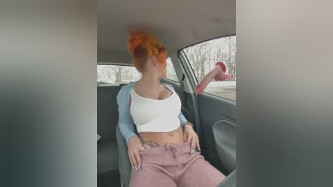 Media: Video of a woman with vibrant orange hair in a messy bun, wearing a white crop top and pink sweatpants, sitting in a car.