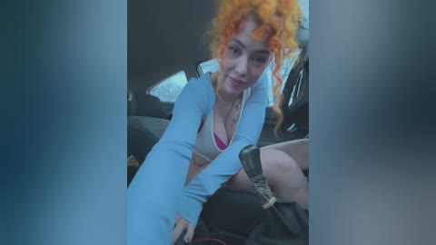 Media: A video of a woman with curly orange hair and light skin, wearing a light blue long-sleeved top, pink bra, and black shorts, sitting in a car.