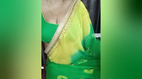 Media: A video of a woman in a green and yellow saree with a gold-embellished border, wearing a gold necklace, seated in a dimly lit room with blurred background.