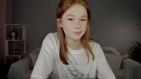 Media: Video of a young Caucasian girl with long red hair, fair skin, and light makeup. She wears a white t-shirt with a graphic design, sitting on a beige couch in a dimly lit living room.