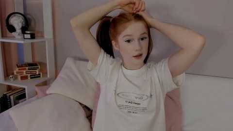 Media: A video of a pale-skinned woman with long red hair in a ponytail, wearing a white T-shirt with \"TAYLOR\" written in black. She's stretching on a white couch in a cozy room with shelves and a clock in the background.