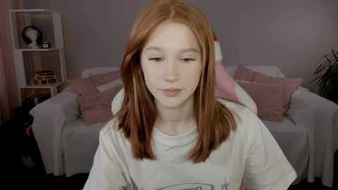 Media: Video of a young, fair-skinned girl with long, straight red hair, wearing a gray T-shirt, sitting in a cozy, light-pink and white bedroom with a sofa, shelves, and a plant.