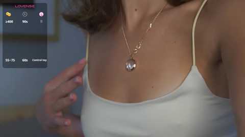 Media: A close-up video of a woman in a white spaghetti-strap tank top, wearing a gold necklace with a heart pendant, indoors. The background is blurred.