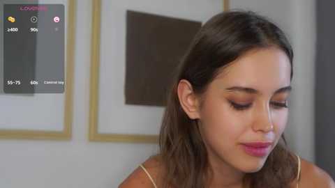 Media: Video of a young Latina woman with long, wavy dark hair, wearing pink lipstick, looking down, in an indoor setting with framed art in the background.