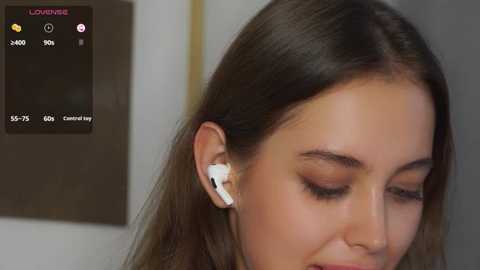 Media: A close-up video of a young woman with light skin and brown hair, wearing white earbuds, listening to music. She has a calm, focused expression. The background is blurred, featuring a white wall.