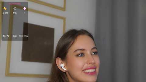 Media: A video of a smiling young woman with light skin and brown hair, wearing white AirPods, against a blurred background with gold-framed art.