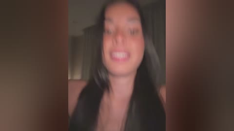 Media: Video of a young woman with long black hair and light skin, smiling widely, wearing a black top, blurred background.