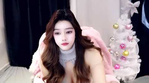 Media: Video of a young East Asian woman with long, wavy brown hair, wearing a gray turtleneck sweater. She sits in front of a white, pink, and gold Christmas tree.