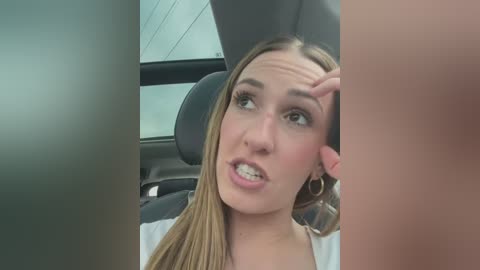 Media: Video of a young woman with long blonde hair and braces, sitting in a car. She has a surprised expression and a hand on her cheek. The car's interior is visible, with a window and gray seats.