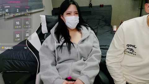 Media: Video of an Asian woman with long black hair in a gray hoodie and face mask, sitting on a black chair in a hospital room with a TV showing a schedule and a person in a white jacket.