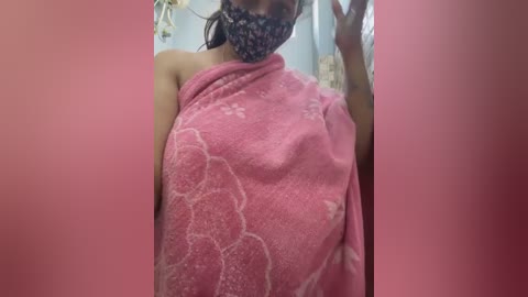 Media: Video of a woman with medium skin tone, wearing a pink towel with white floral patterns, a floral face mask, and dark hair, in a bathroom.