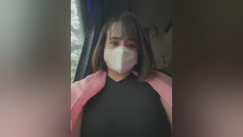 Media: A video of an Asian woman with shoulder-length dark hair, wearing a white face mask, black top, and pink jacket, sitting indoors near a window with greenery outside.