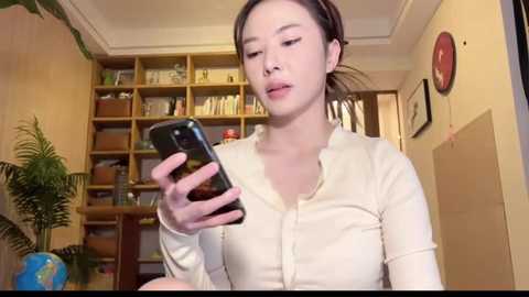 Media: Video of an Asian woman with fair skin, long black hair tied back, wearing a cream blouse, taking a selfie in a cozy, book-filled room with a wooden bookshelf and framed art.