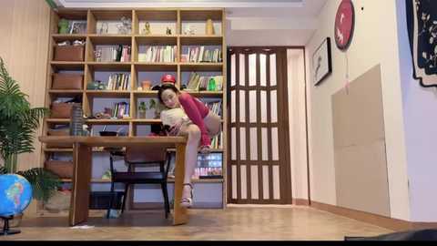 Media: A video of an Asian woman in a red shirt, wearing glasses, sitting at a wooden desk with a laptop, surrounded by a bookshelf filled with books, in a cozy, brightly lit room with a globe and framed art on the walls.