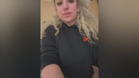 Media: A video of a woman with long, curly blonde hair, wearing a black hoodie with a small red logo, standing in a dimly lit, blurred background.