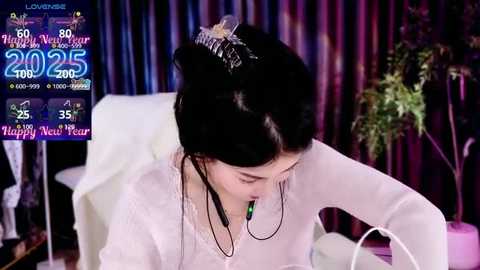 Media: Video of a woman with black hair wearing a tiara and headset, seated at a white desk, with a calendar and plant in the background.