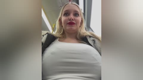 Media: Video of a blonde woman with fair skin and red lipstick, wearing a white tank top and black jacket, sitting in a subway car, blurred faces in the foreground.