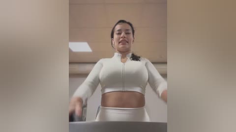 Media: Video of a Latina woman with medium skin tone and medium build, wearing a white, long-sleeved crop top and high-waisted leggings, smiling while running on a treadmill in a gym.