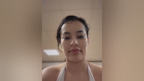 Media: Video of a young woman with light brown skin and dark hair pulled back, wearing a white halter top, standing in a beige-tiled corridor with fluorescent lighting.