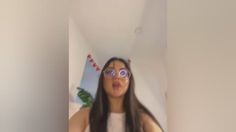 Media: Video of a young woman with long black hair, wearing large glasses and a white tank top, making a kissing face. She stands in a hallway with a green plant and heart decorations on the wall in the background.