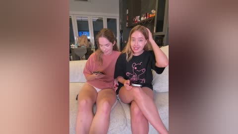 Media: Video of two young women with fair skin, sitting on a white couch in a modern living room. They wear casual clothes and are engrossed in their smartphones, with the background showing a TV and shelves with toys.