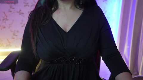 Media: Video of a woman with long dark hair, wearing a black V-neck dress with a belt and three-quarter sleeves, seated in a dimly lit room with a floral-patterned wall and blue lighting.