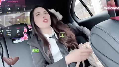 Media: Video of a young woman with long brown hair, wearing a gray blazer and white shirt, holding a tissue, inside a car, with a blurred cityscape and digital screens in the background.