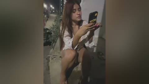 Media: Video of a young Latina woman with long brown hair, wearing a white floral blouse and black shorts, sitting on a ledge at night, taking a selfie with a black phone.